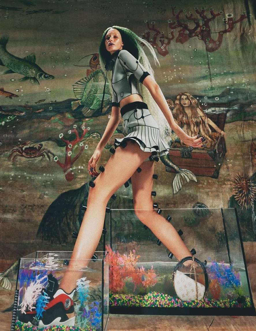A fashion model in an aquatic-themed editorial shot, wearing a grey two-piece outfit while standing in small, water-filled fish tanks.
