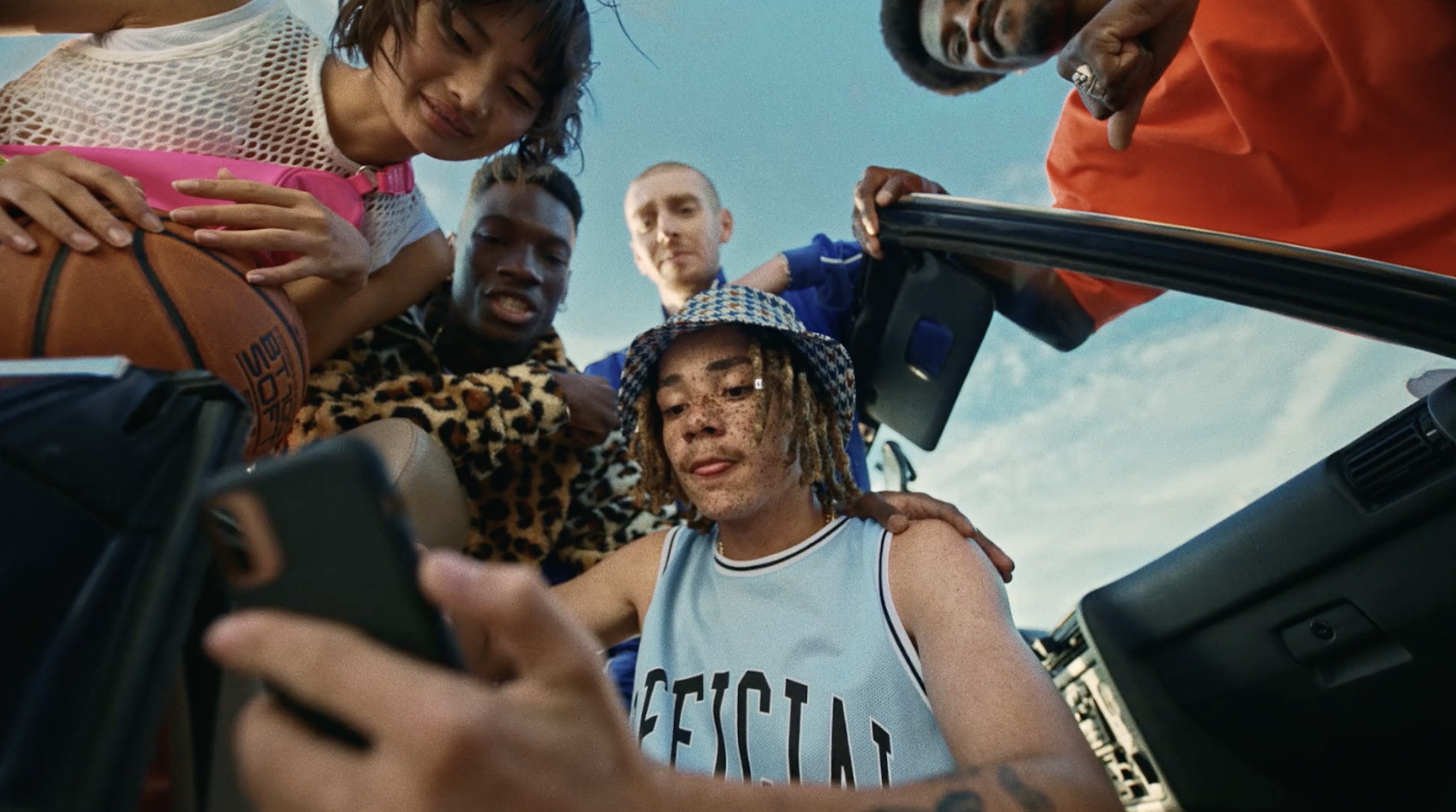Low-angle view of six diverse young adults posing with a basketball and cell phone outside a car under bright, sunny skies.