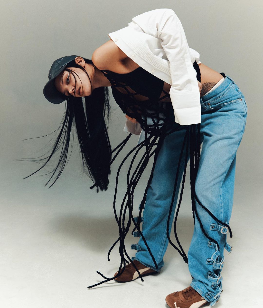 Model in blue denim jeans, black string top, white jacket, and cap bending over against a grey background