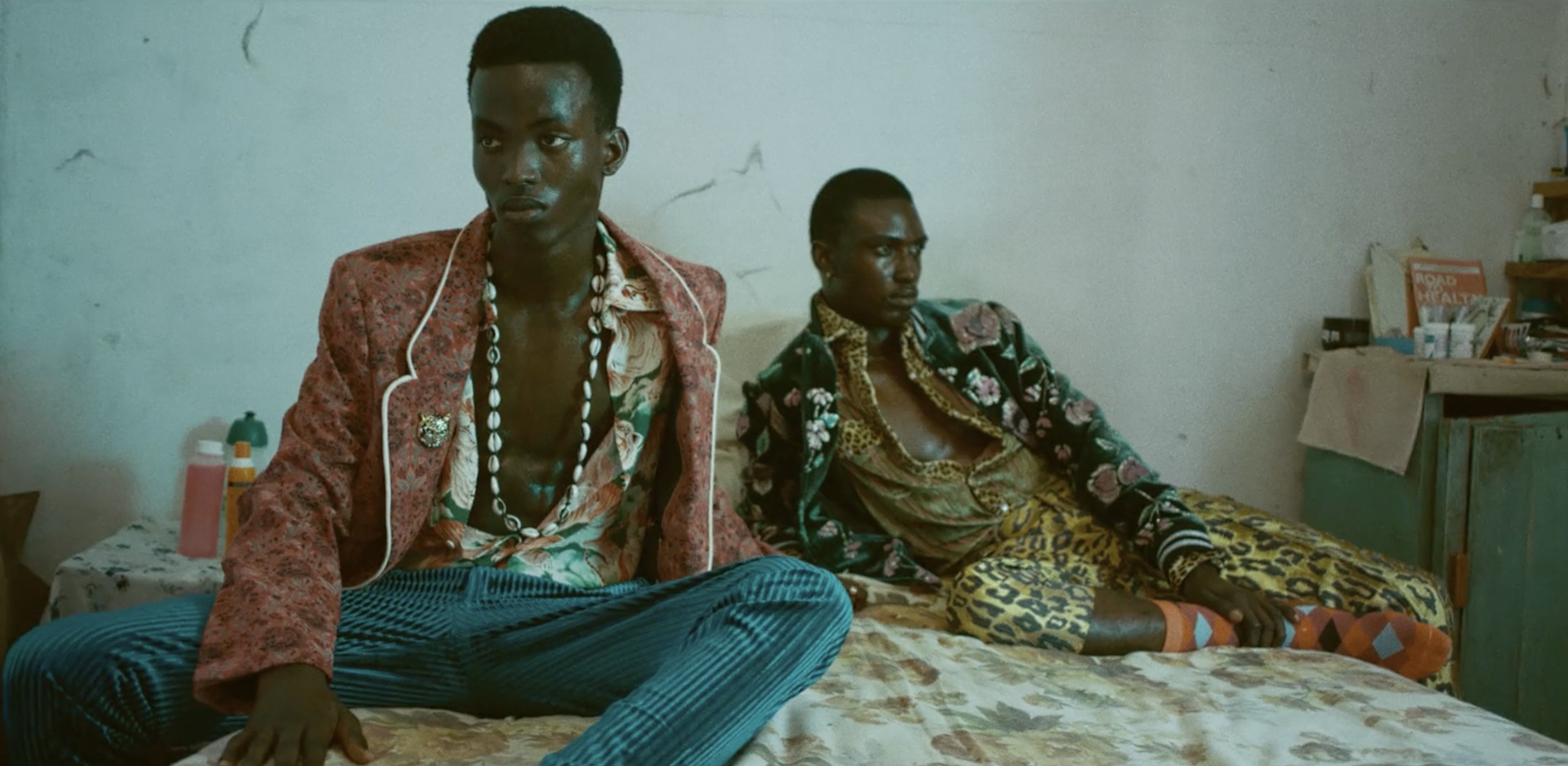 Two dark-skinned adult males sit on a bed wearing fashionable blazers and shirts with animal prints in a vintage-style indoor setting.