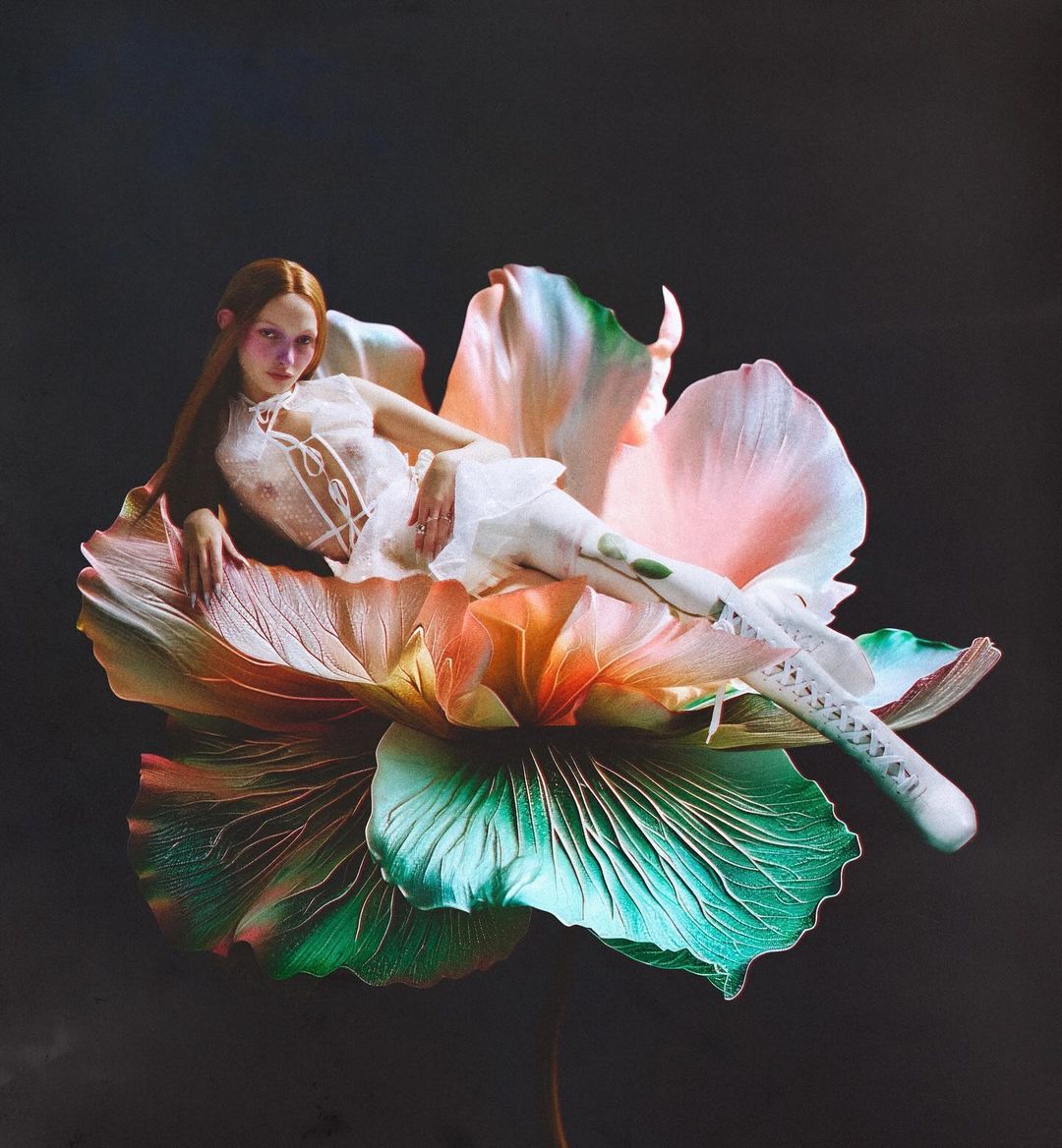Woman in frilly white dress and boots lies on a giant flower with pink, green and orange petals against a dark grey background.