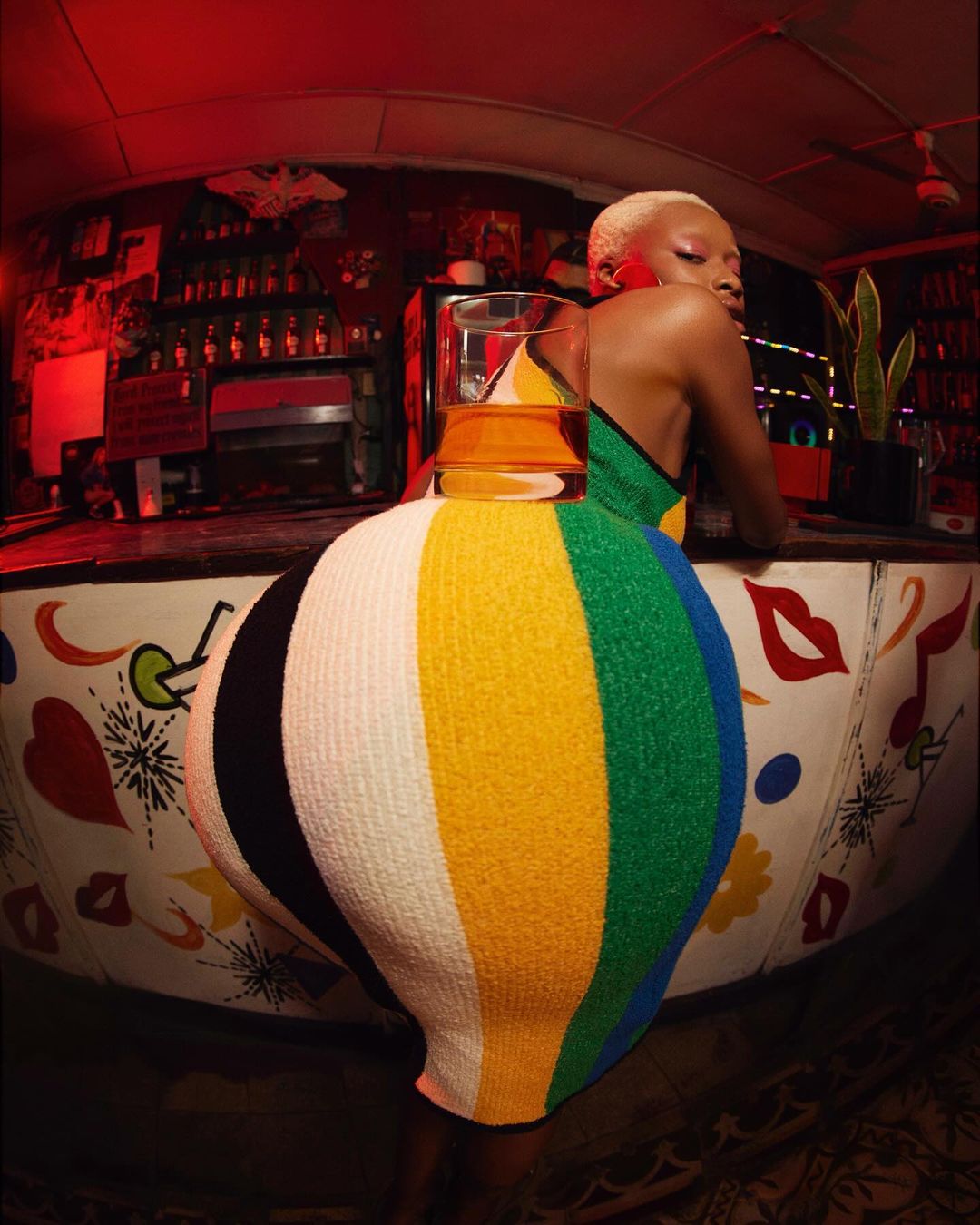 Woman in a colorful dress stands behind a decorated bar, posing with a drink in a glass in a dimly lit bar, fisheye lens.