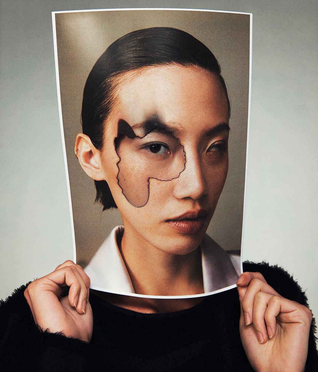 Conceptual portrait with a woman holding a photo cutout revealing part of her face, set against a grey background, fashion-forward presentation.