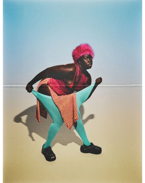 Fashionable dark-skinned adult female in pink fur hat posing outdoors in bright blue tights and black platform shoes with a gradient backdrop.