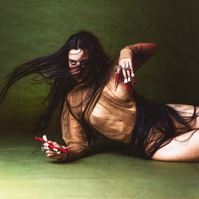 Brunette woman with long hair poses on a green surface, wearing sheer top and long red and white pointed nails, looking at the camera.