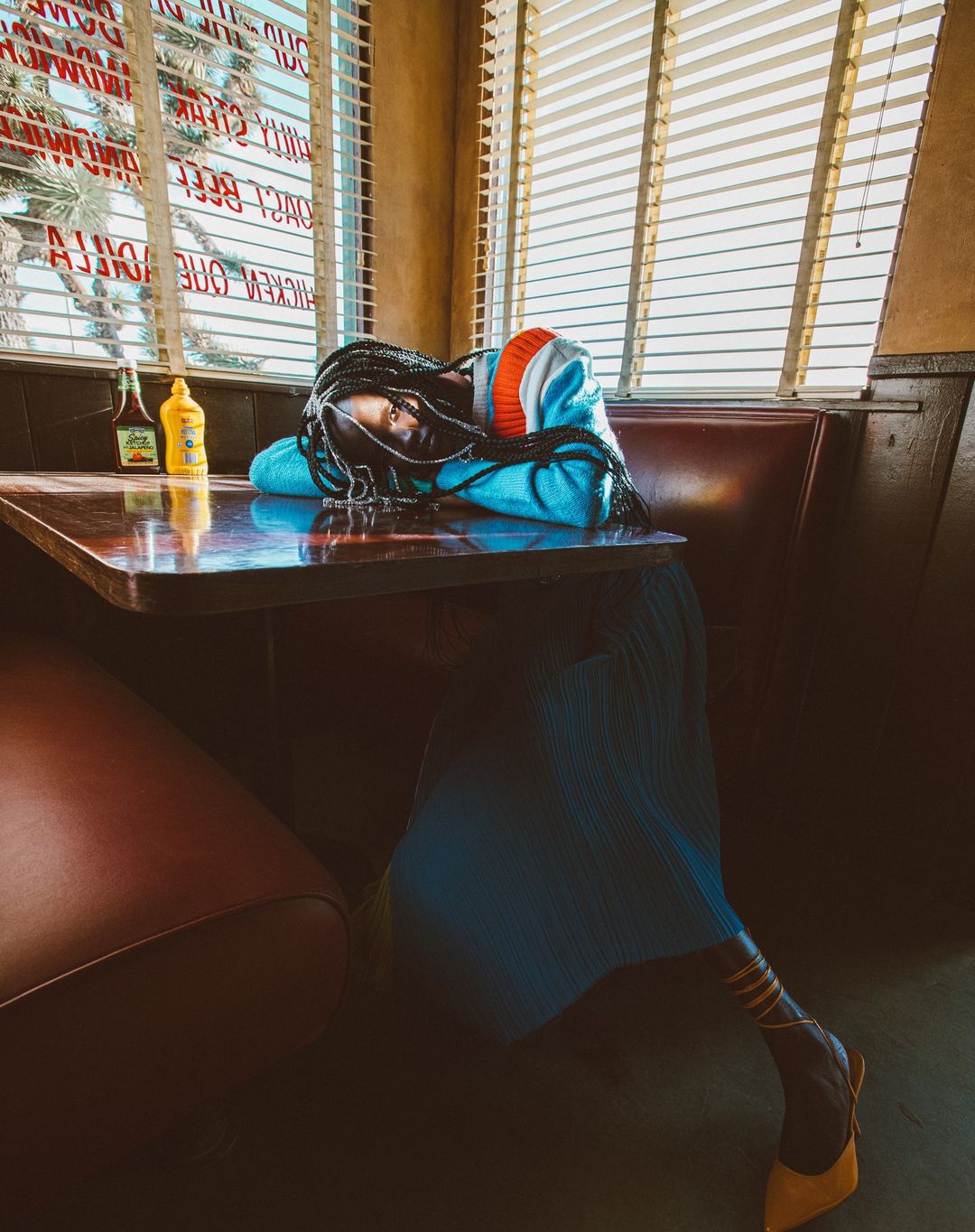 Woman rests head on table in vintage diner. Braided hair, colorful sweater, pleated skirt, and high heels. Mustard and hot sauce on table.