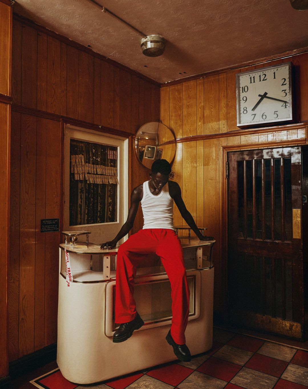 Stylish Black man sits on a vintage counter in red pants and white tank top in retro room with wood paneling, clock, and checkered floor.