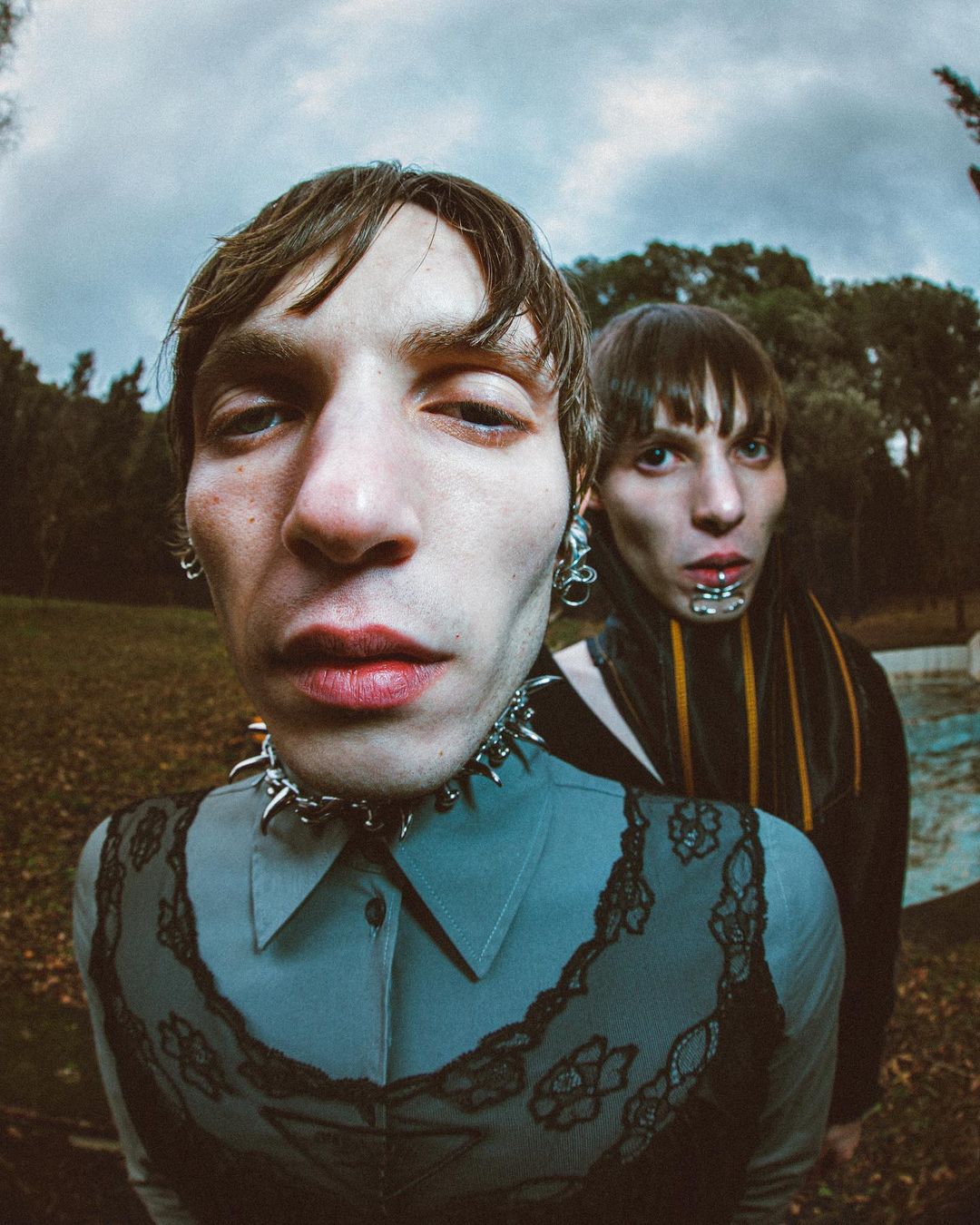 Two young adults pose in an outdoor portrait. One person wears a spiked collar and lace-trimmed top; the other wears silver lip jewelry.