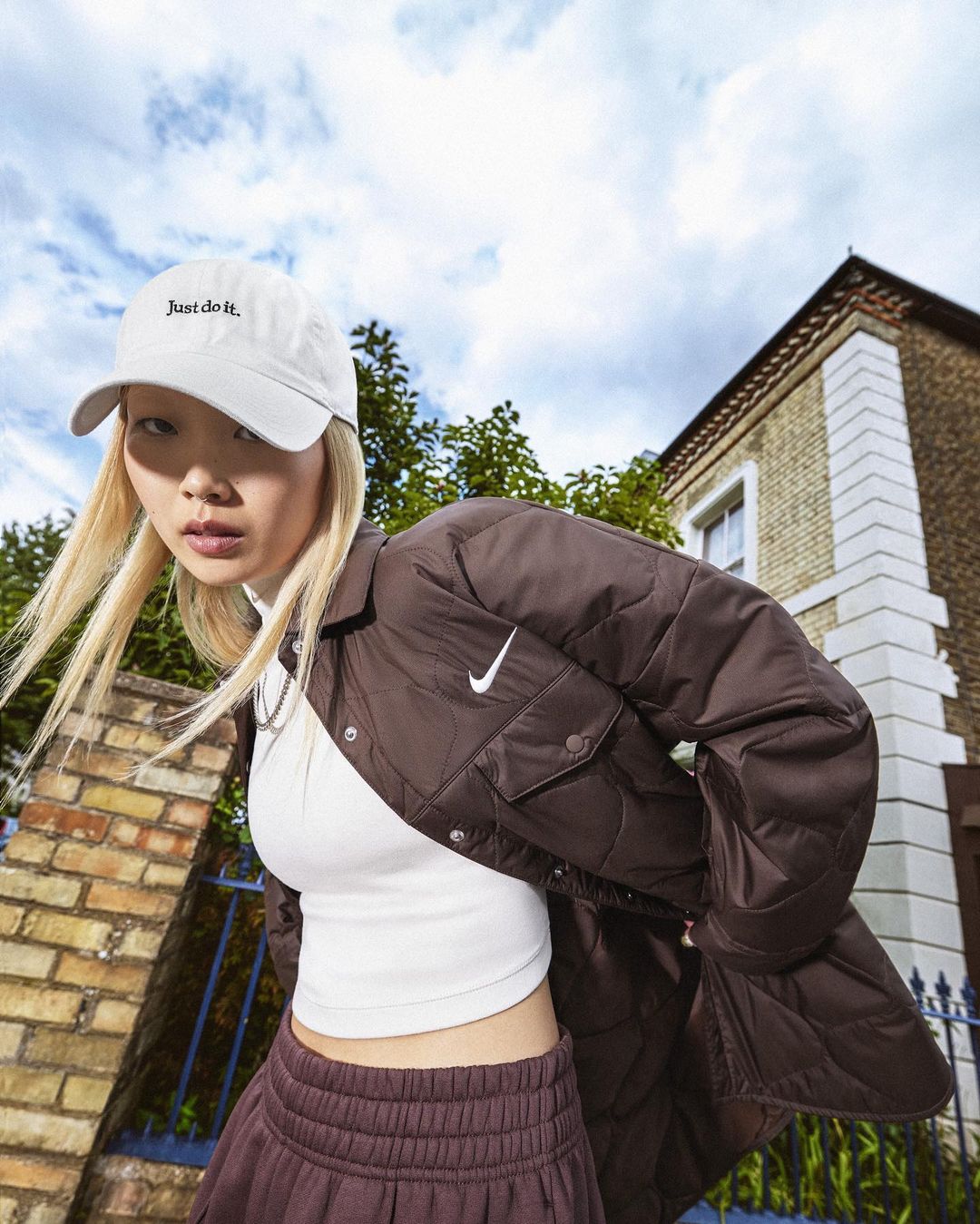 A young Asian woman models Nike activewear with a white logo cap, brown jacket, white top, and maroon pants against a brick building backdrop.