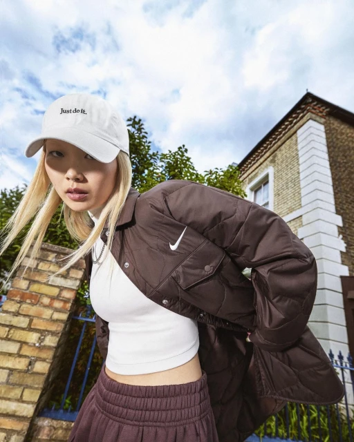 A young Asian woman models Nike activewear with a white logo cap, brown jacket, white top, and maroon pants against a brick building backdrop.