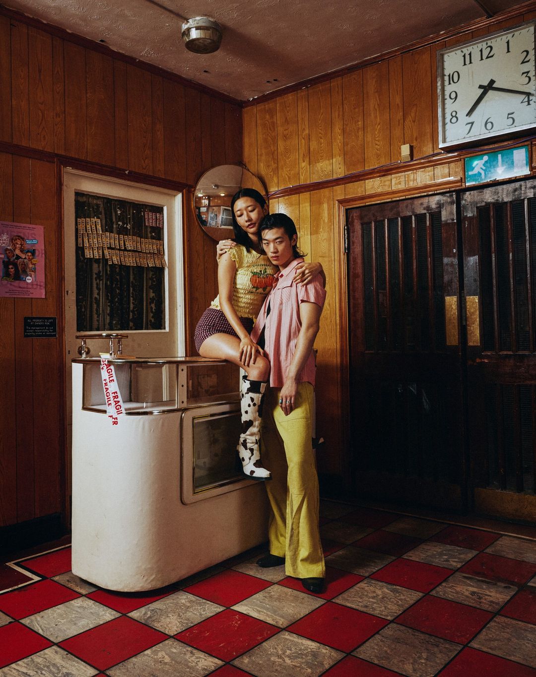 Stylish couple in vintage-themed diner: Asian woman sits on counter, man stands beside in flared yellow pants, red checkered floor.