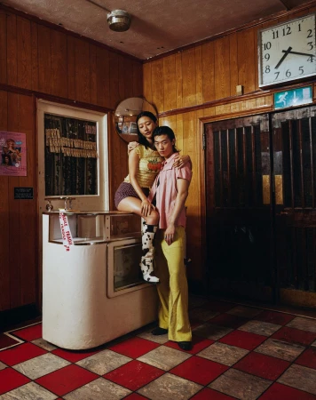 Stylish couple in vintage-themed diner: Asian woman sits on counter, man stands beside in flared yellow pants, red checkered floor.