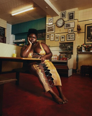 Plus-size model poses indoors in a yellow dress with open lattice detail, seated at a wooden table in an eatery with tiled walls.