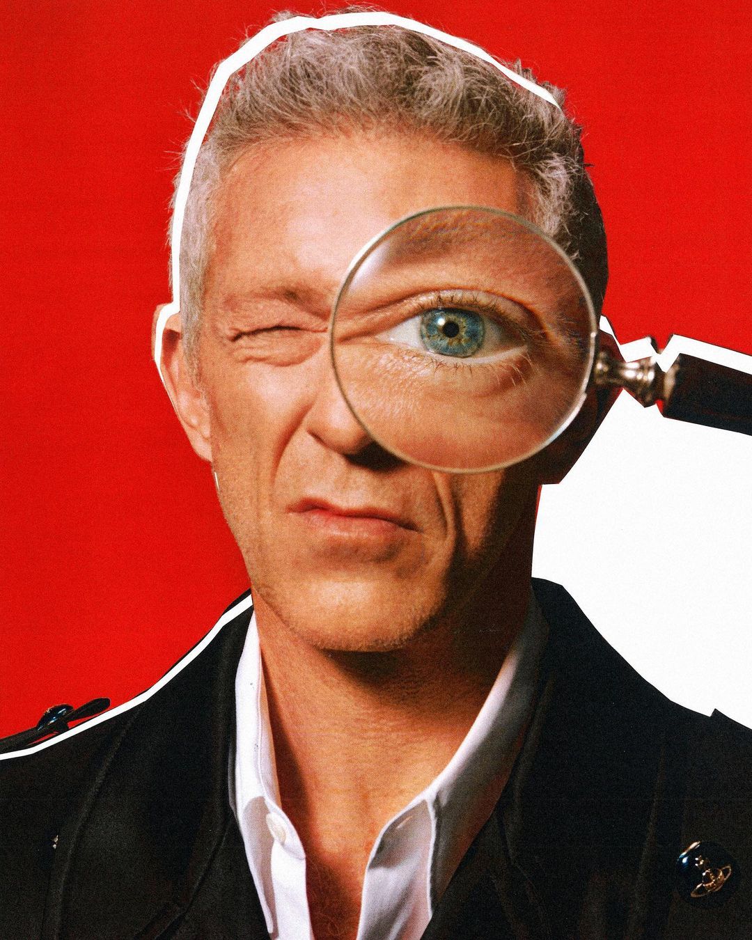 Close-up portrait of a grey-haired man winking with a magnifying glass held to his eye, set against a red and white background.