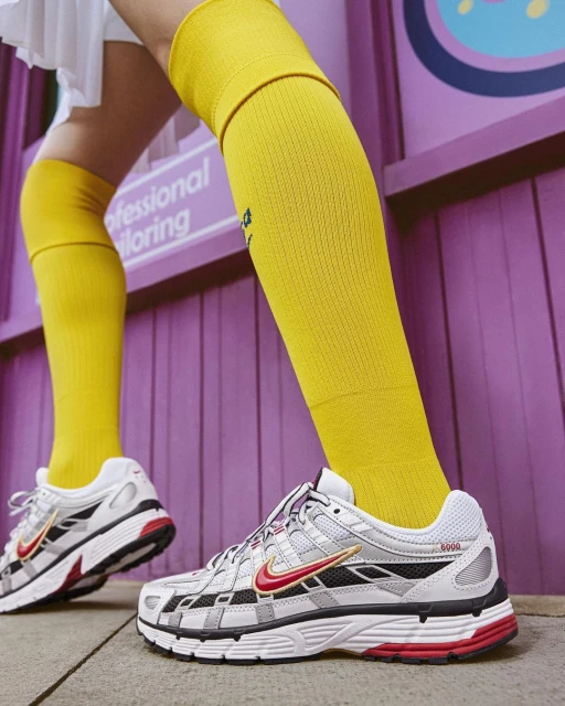 Close-up of white and grey Nike sneakers with red swoosh, worn with yellow socks. Shot in a studio against a purple wall.