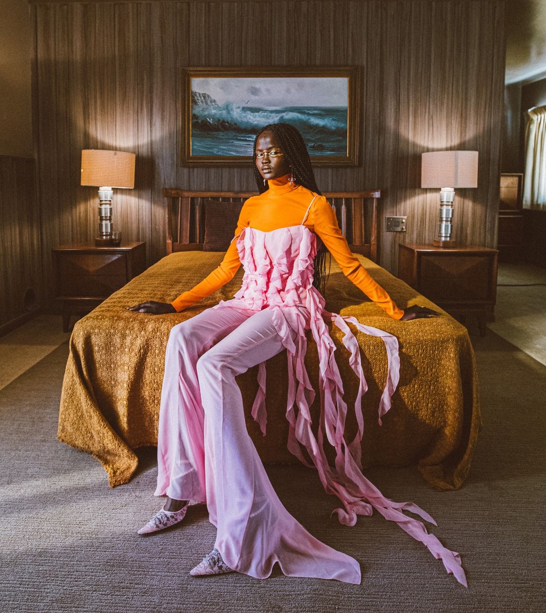 Elegant woman in orange top and pink pants posing on a bed in a warmly lit room with a painting of a seascape on the wall.
