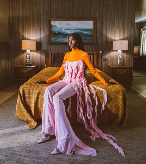 Elegant woman in orange top and pink pants posing on a bed in a warmly lit room with a painting of a seascape on the wall.