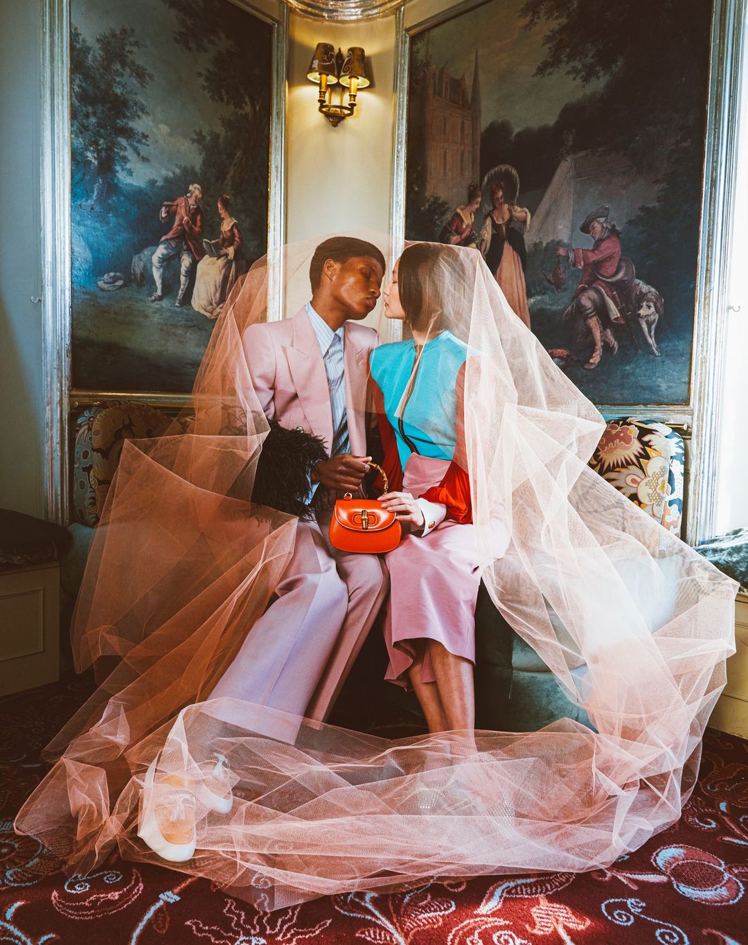A romantic indoor portrait captures an African-American man and an Asian woman sitting closely, draped in peach tulle with a painted backdrop.