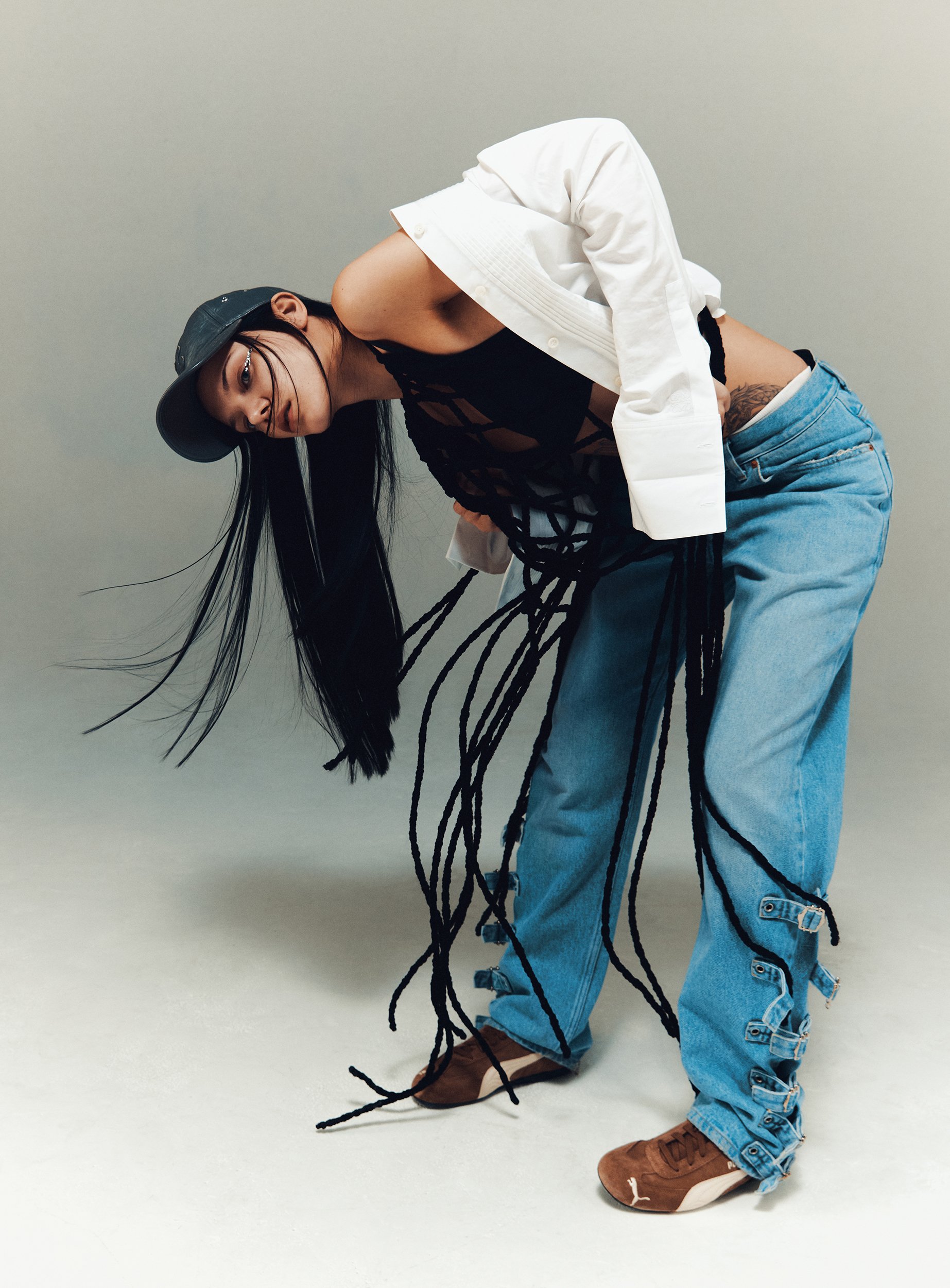 Woman in jeans and a top poses with long black hair and a baseball cap against a neutral background in a studio fashion shot.