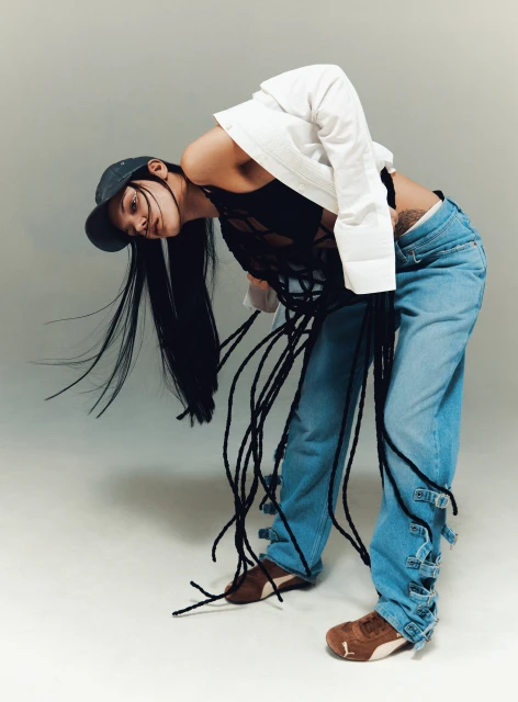 Woman in jeans and a top poses with long black hair and a baseball cap against a neutral background in a studio fashion shot.