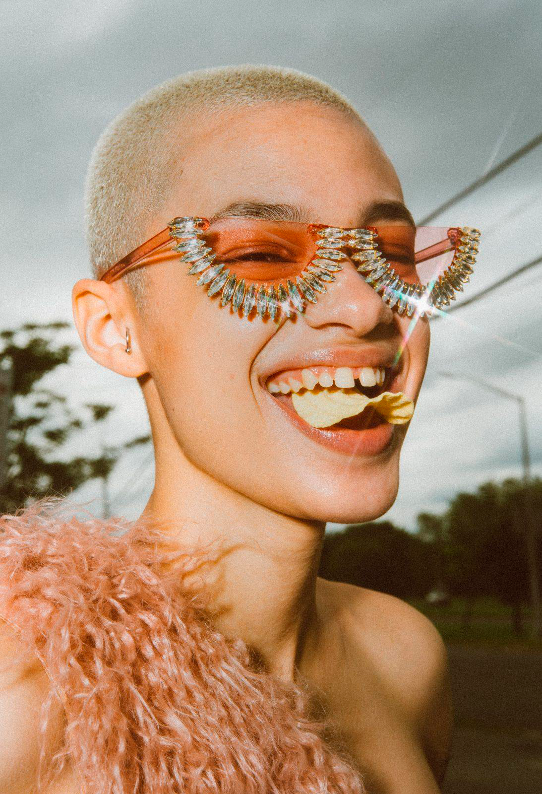 Smiling person with buzzcut poses outdoors wearing novelty sunglasses, shoulder visible with fuzzy light pink fabric and eating potato chips.