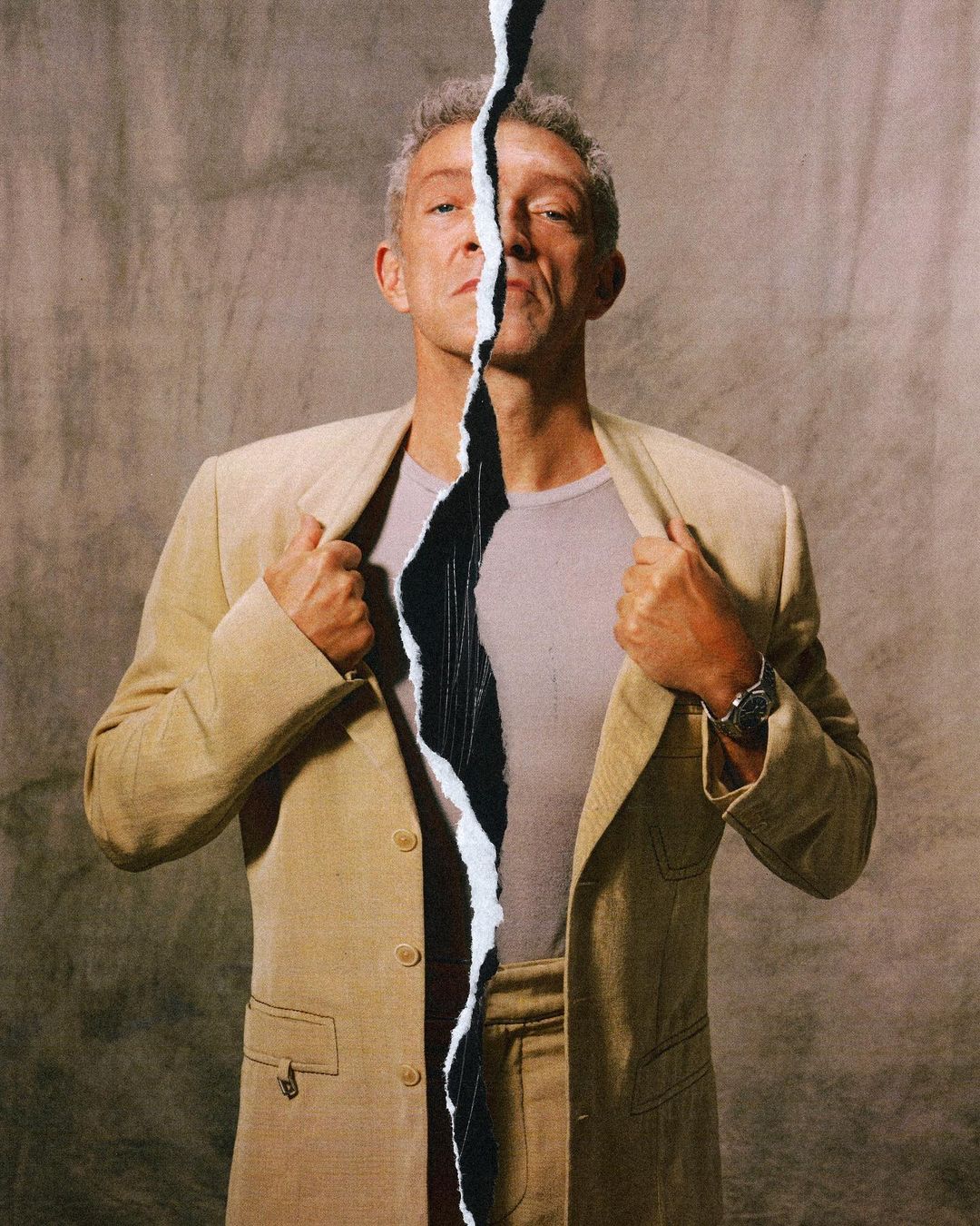 Mature adult male in beige suit and t-shirt with torn paper effect in front of gray background, looking at the camera. 