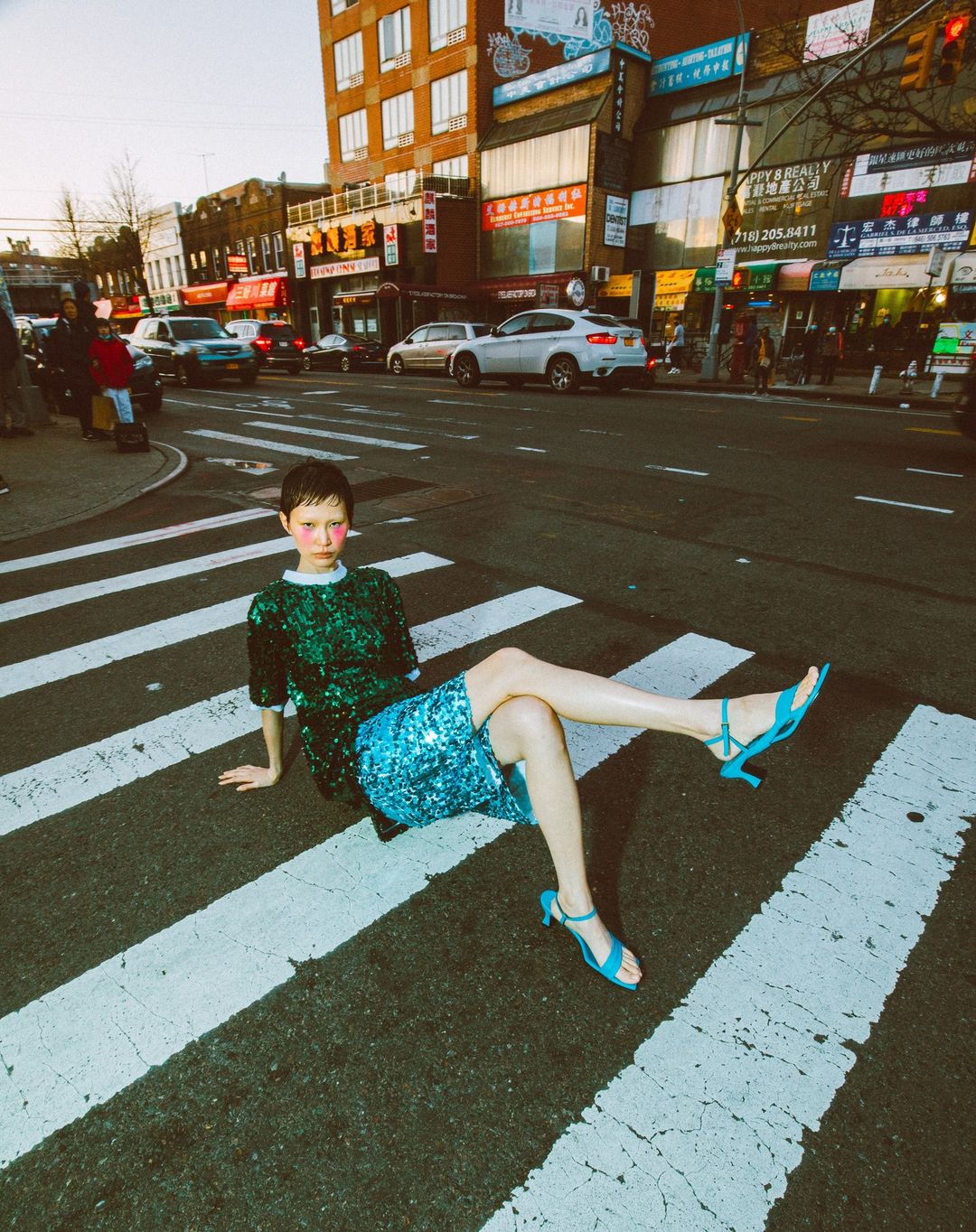 A woman in a sequin dress and blue heels sits in a crosswalk on a city street with buildings and cars in the background, captured at eye-level.