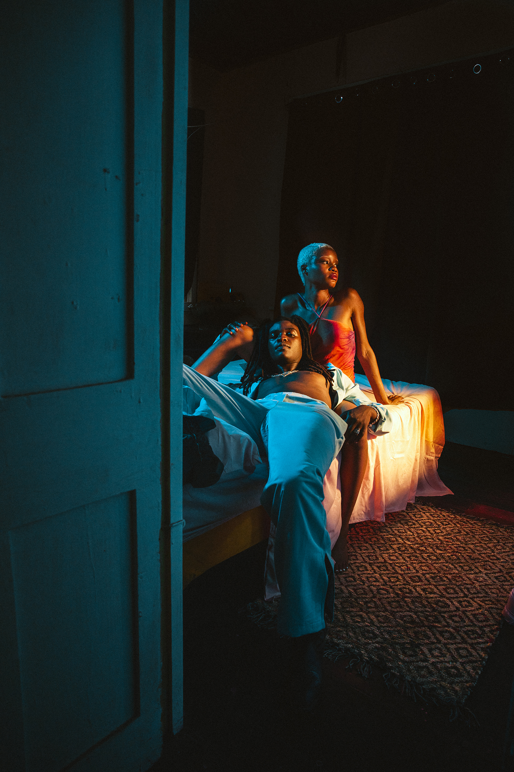 Two people in a dimly lit room, an adult male laying in bed and an adult female sitting behind. The scene has warm and cool lighting.