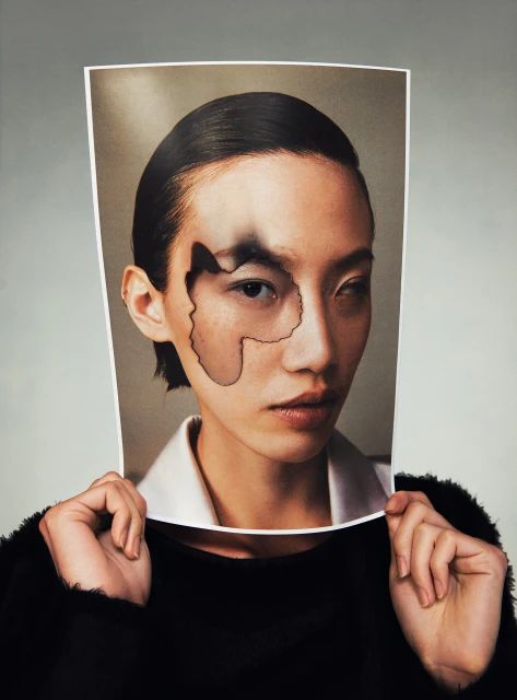 Asian woman holds up a picture of her face with dark irregular shapes on it in front of her face in a studio setting.
