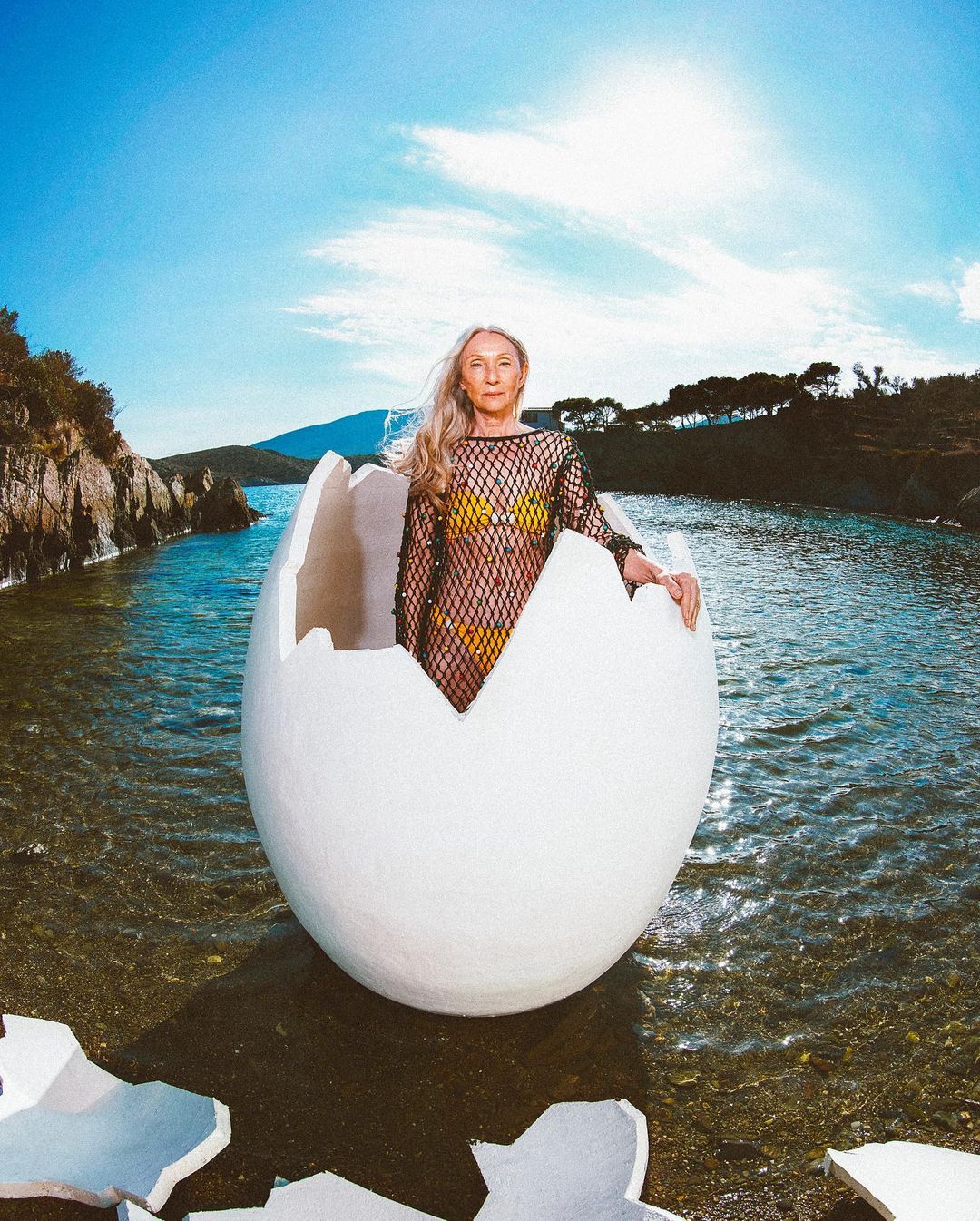 Woman in a net dress poses inside a cracked egg in shallow water against a sky and ocean backdrop with rocky hills in the distance.