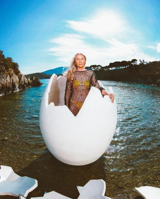 Woman in a net dress poses inside a cracked egg in shallow water against a sky and ocean backdrop with rocky hills in the distance.
