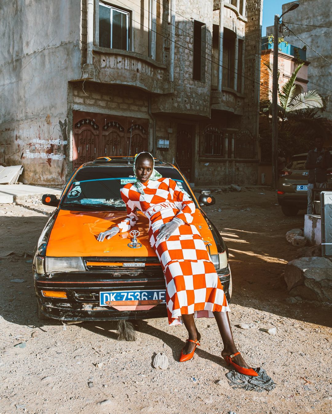 A woman in a checkered dress leans against an orange taxi on a dirt road in front of weathered buildings in an urban setting.