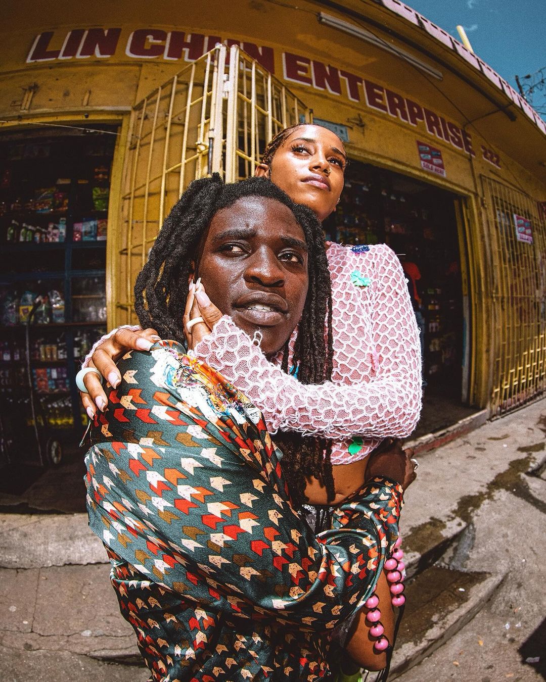 Two people pose in front of a shop: one with dreadlocks and a patterned shirt, the other with a pink top, in a sunny outdoor setting.