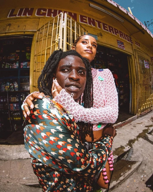 Two people pose in front of a shop: one with dreadlocks and a patterned shirt, the other with a pink top, in a sunny outdoor setting.