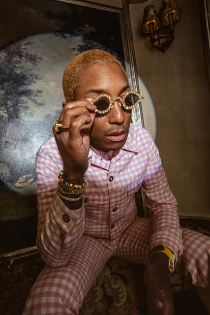 Adult male in pink and white checkerboard suit poses indoors with decorative glasses, gold jewelry, and vintage painting background.