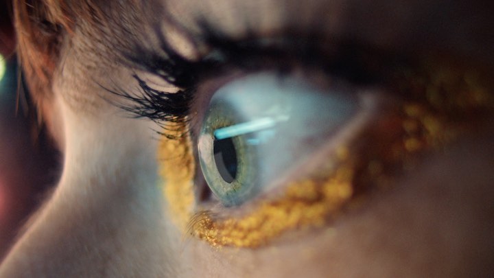 Close-up shot of a green eye with gold makeup on the lower lid and eyelashes. The skin is fair. Indoor lighting.