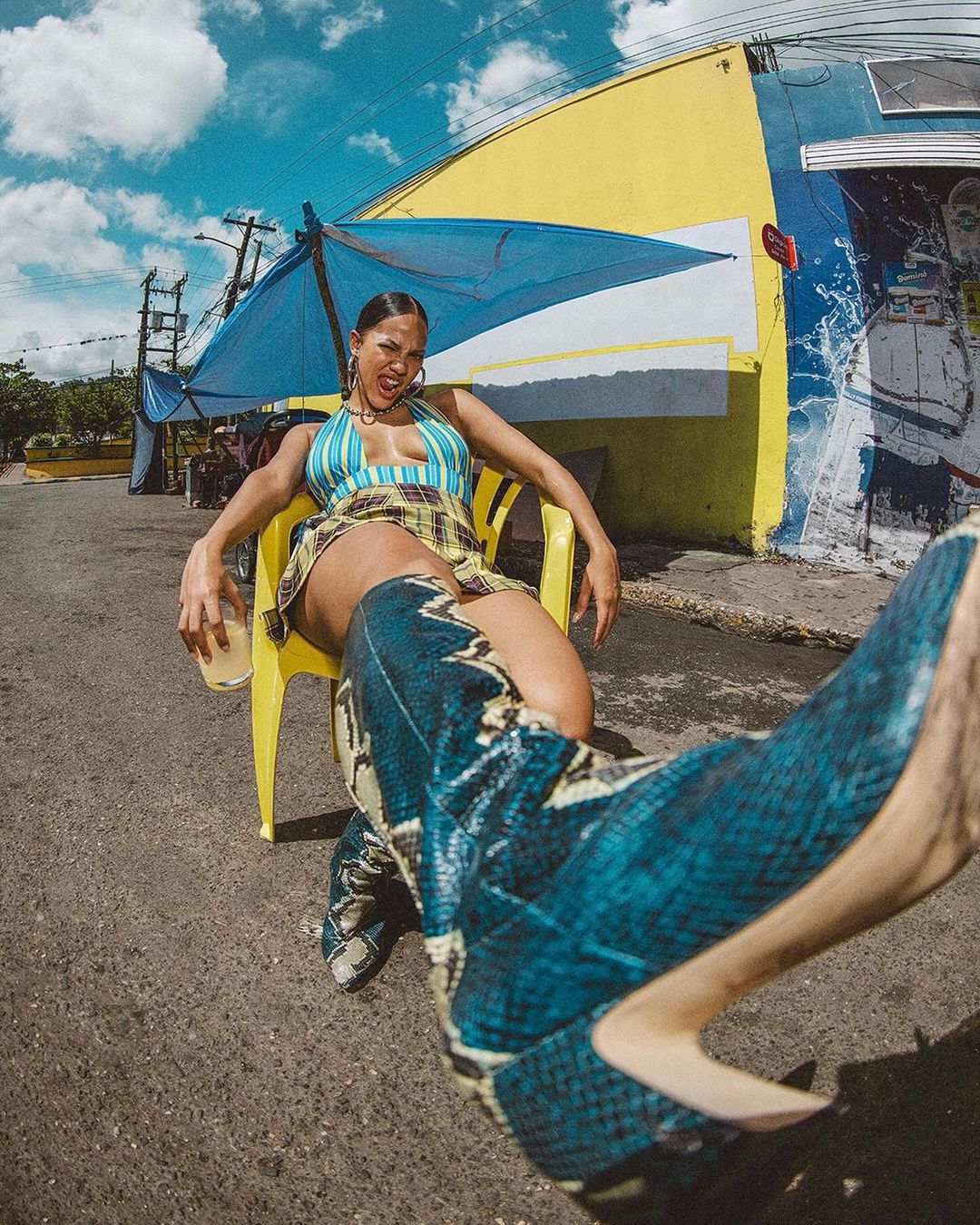 Woman in snakeskin boots and striped halter top holding a drink under a blue parasol by Lou Escobar