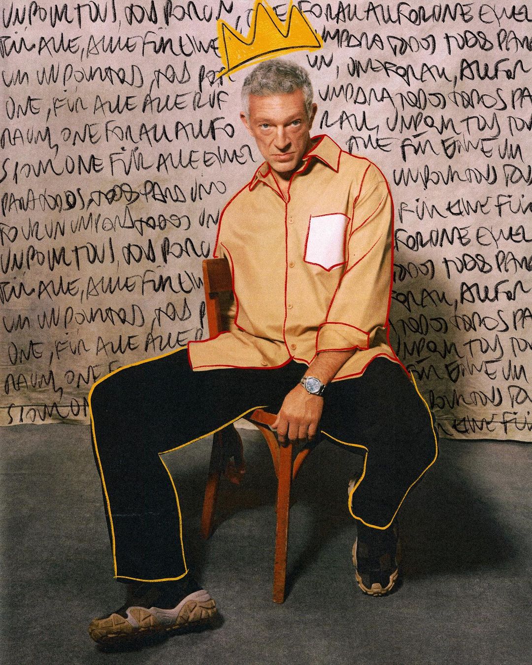 Adult man in a casual button-down shirt and dark pants sitting on a chair in front of a graffiti-covered wall with a drawn crown.