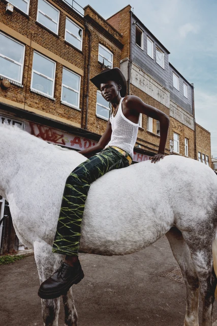 Fashion shot: a black adult male, wearing cowboy hat and green pants, sits on a white horse in an urban setting with brick buildings.