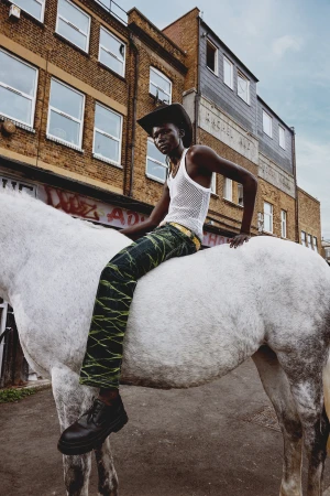 Fashion shot: a black adult male, wearing cowboy hat and green pants, sits on a white horse in an urban setting with brick buildings.