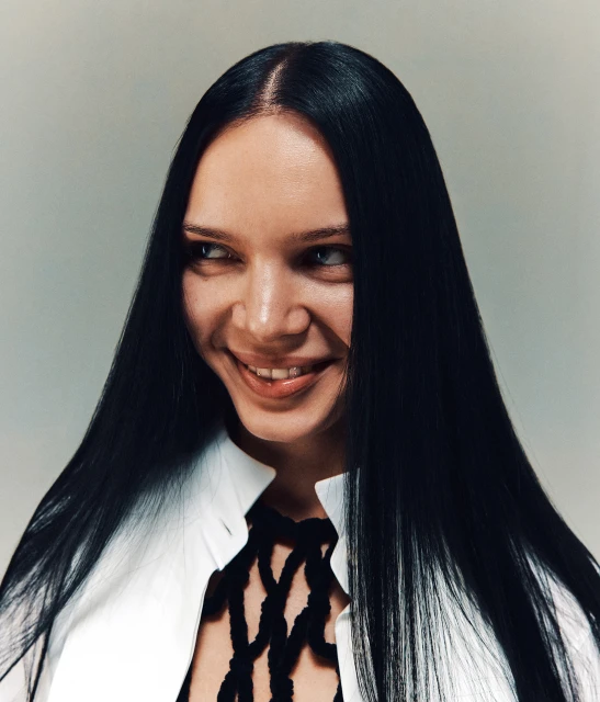 Portrait of a smiling adult woman with long, straight black hair and a white collared shirt against a neutral background in a studio setting.
