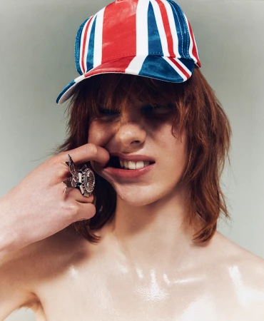 Young adult posing in a studio wearing a British-flag baseball cap and a ring, pulling his mouth open with his fingers.