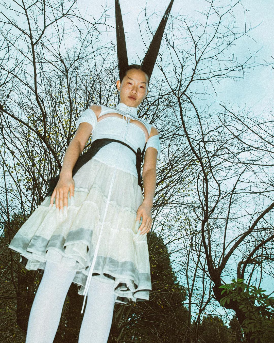 Woman with dramatic hairstyle poses outdoors. She wears a white top, a tiered dress, and white tights, set against trees and a light sky.