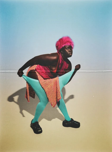 Full body portrait of a dark-skinned adult woman in blue tights, black shoes, and pink hat posing in a studio against a gradient backdrop.