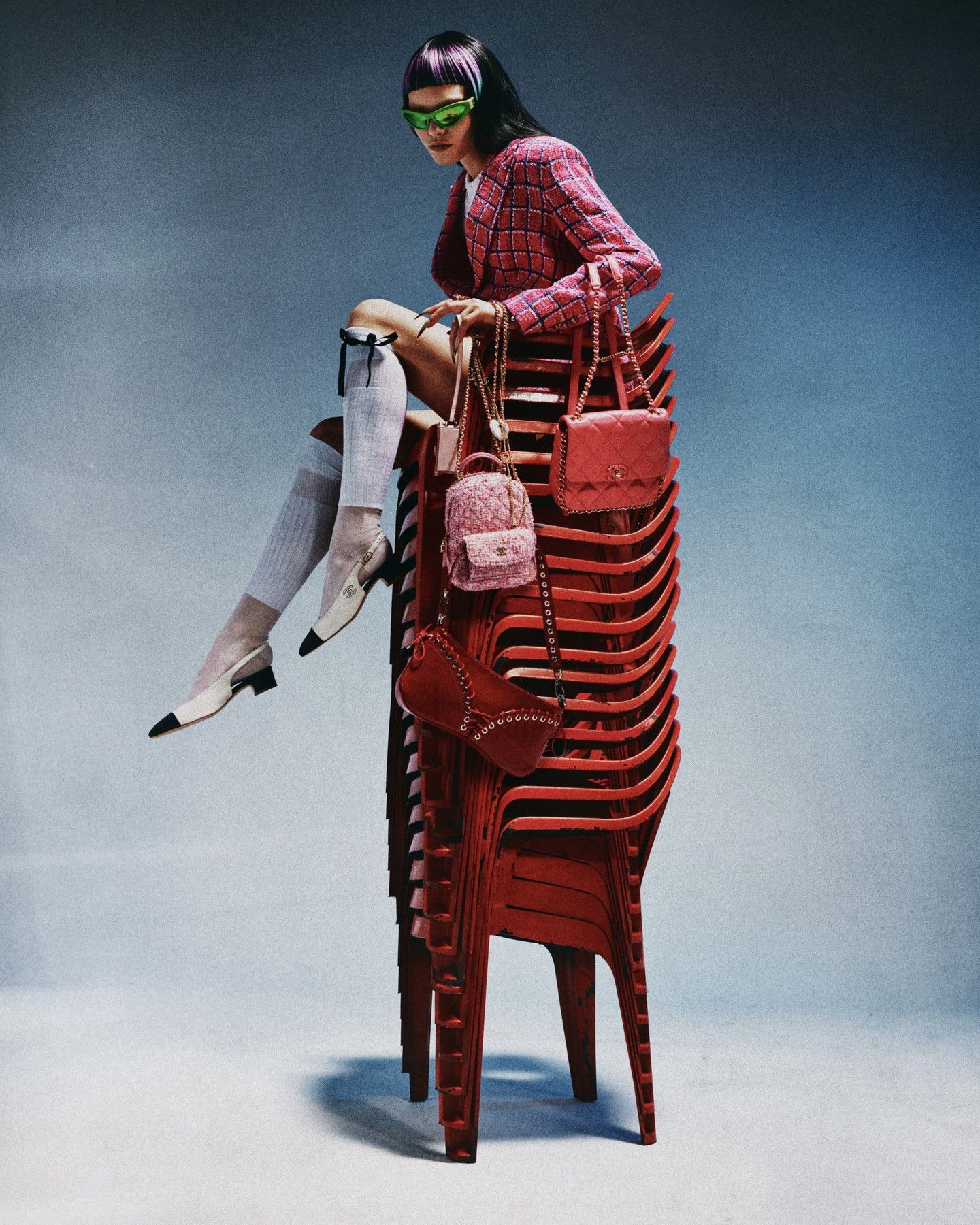 An adult female model with purple-streaked hair sits on a stack of red plastic chairs, holding several purses against a blue gradient background.