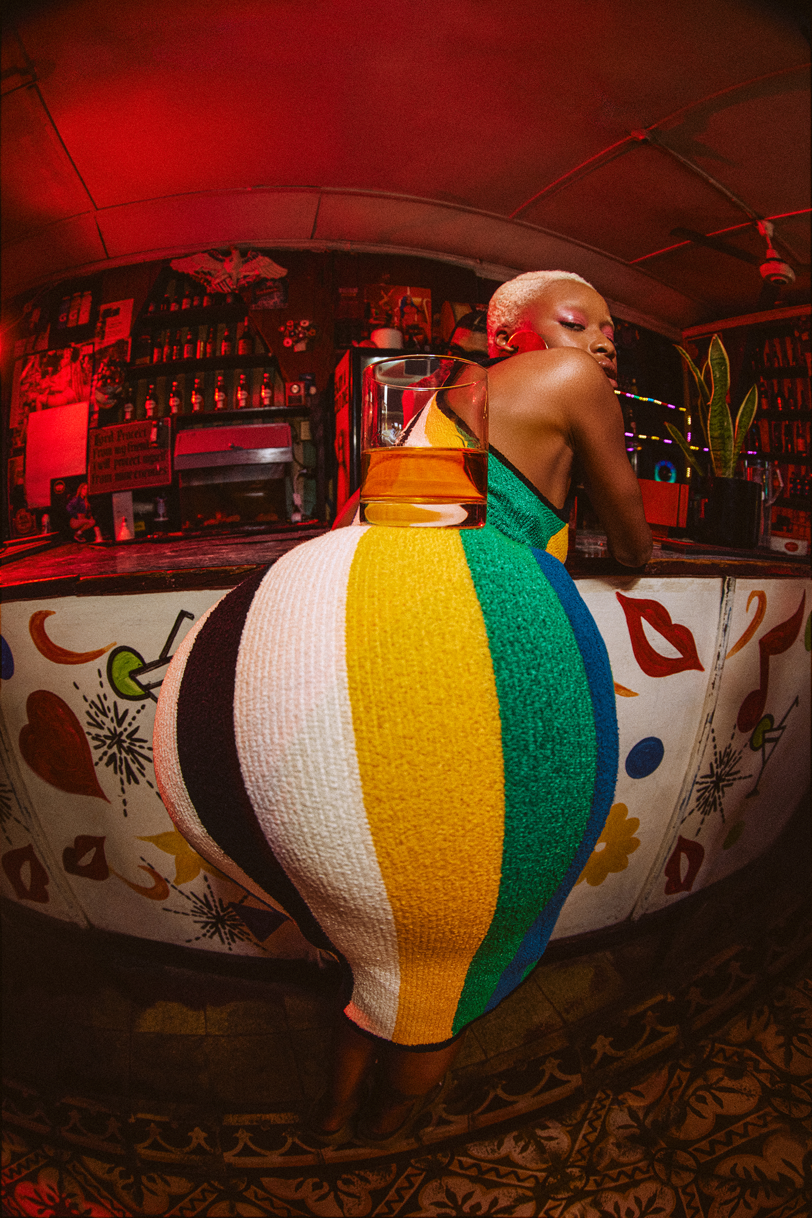 A woman in a colorful striped dress leans on a bar, featuring a cocktail, set indoors under warm red lighting with a fisheye lens effect.