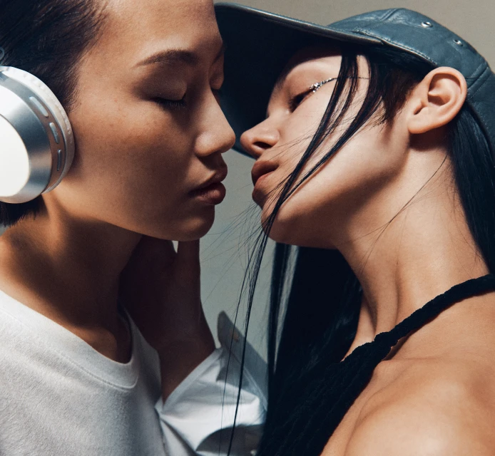 Close-up of two androgynous fashion models with closed eyes facing each other, one with headphones and a white top, the other in a cap.