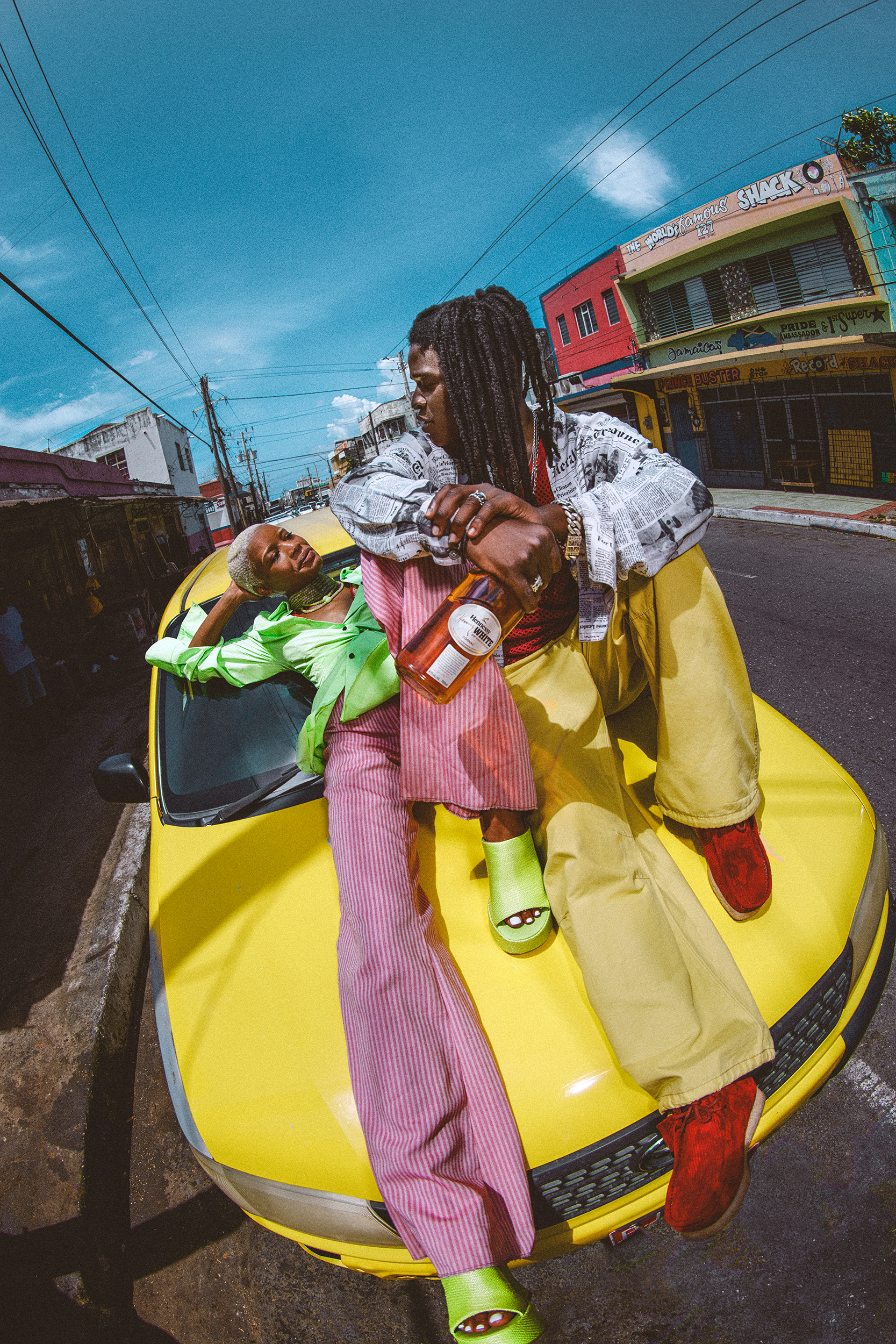 Two stylish people pose on a yellow car in an urban setting, featuring colorful clothing, building exteriors, and a clear blue sky.