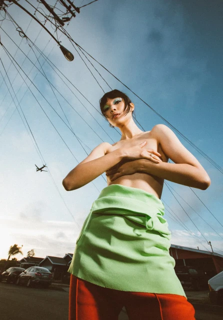 A woman in an outdoor shot, gazing upward at the camera with bright eye makeup, wearing a green skirt and red pants against a sky background.