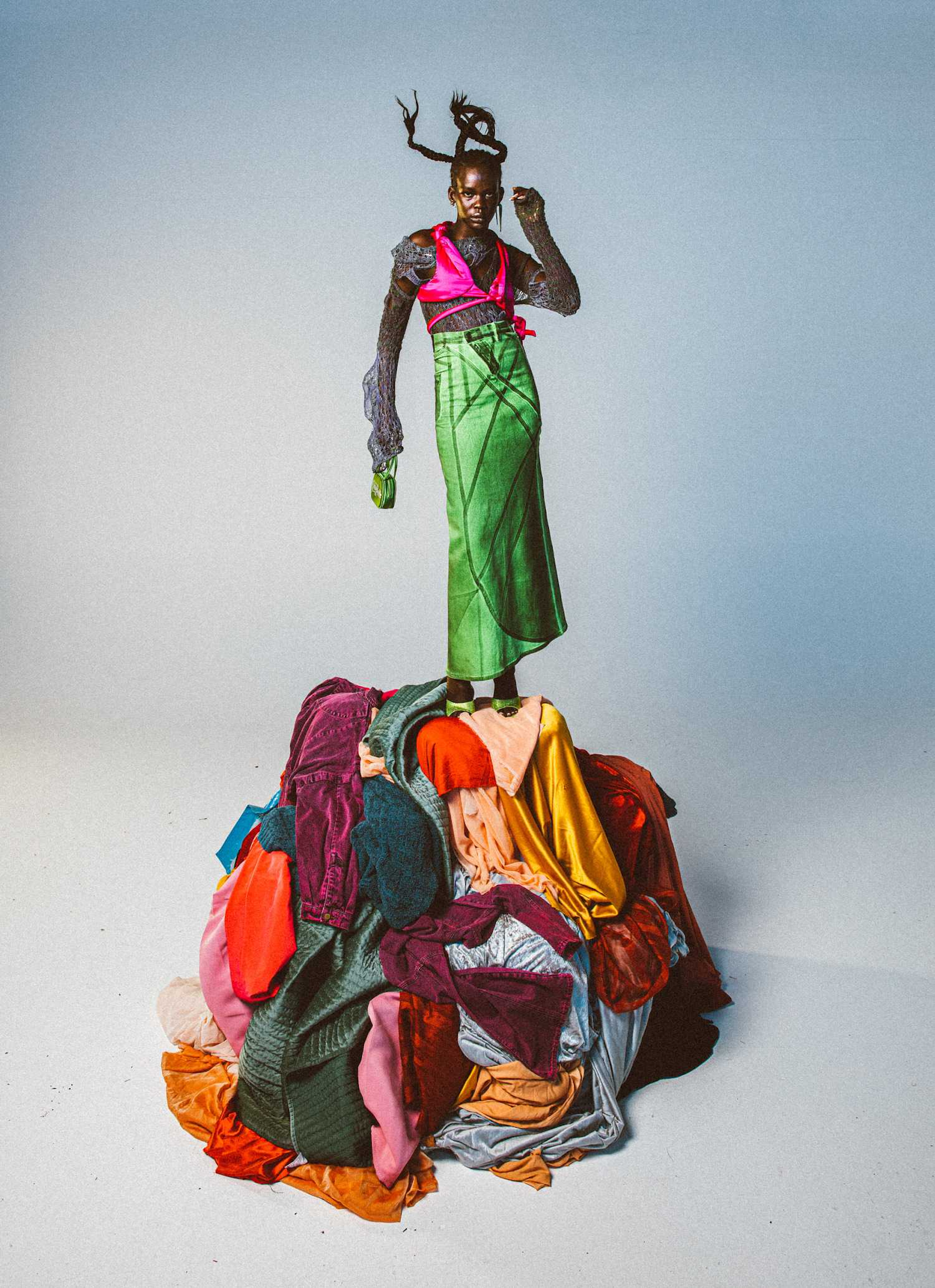 Fashionable black woman stands on pile of colorful clothes in studio wearing green skirt, pink top, patterned sleeves, and holding small purse.